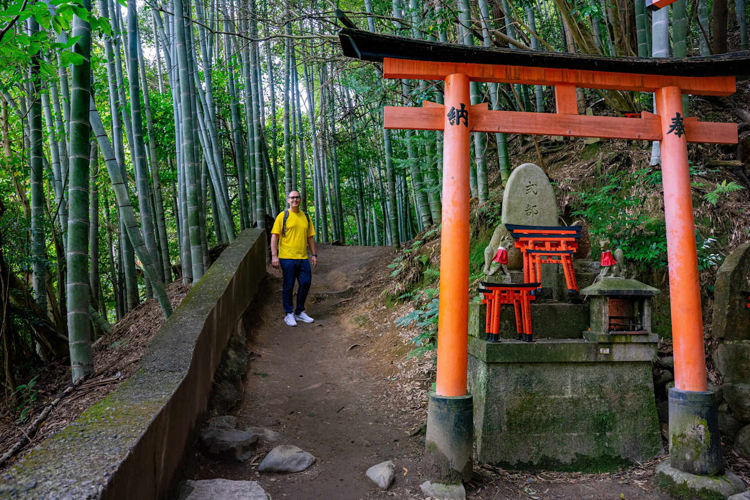 Hiker in yellow standing before red torii gate at Fushimi Inari Shrine surrounded by dense bamboo forest