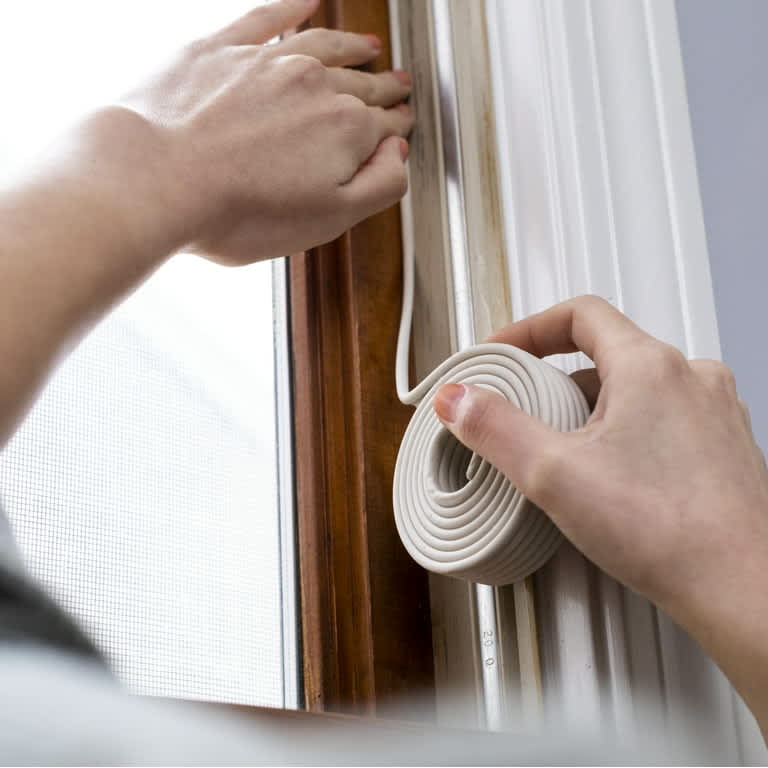 A homeowner using their thumbs to press rope caulk into a drafty window gap