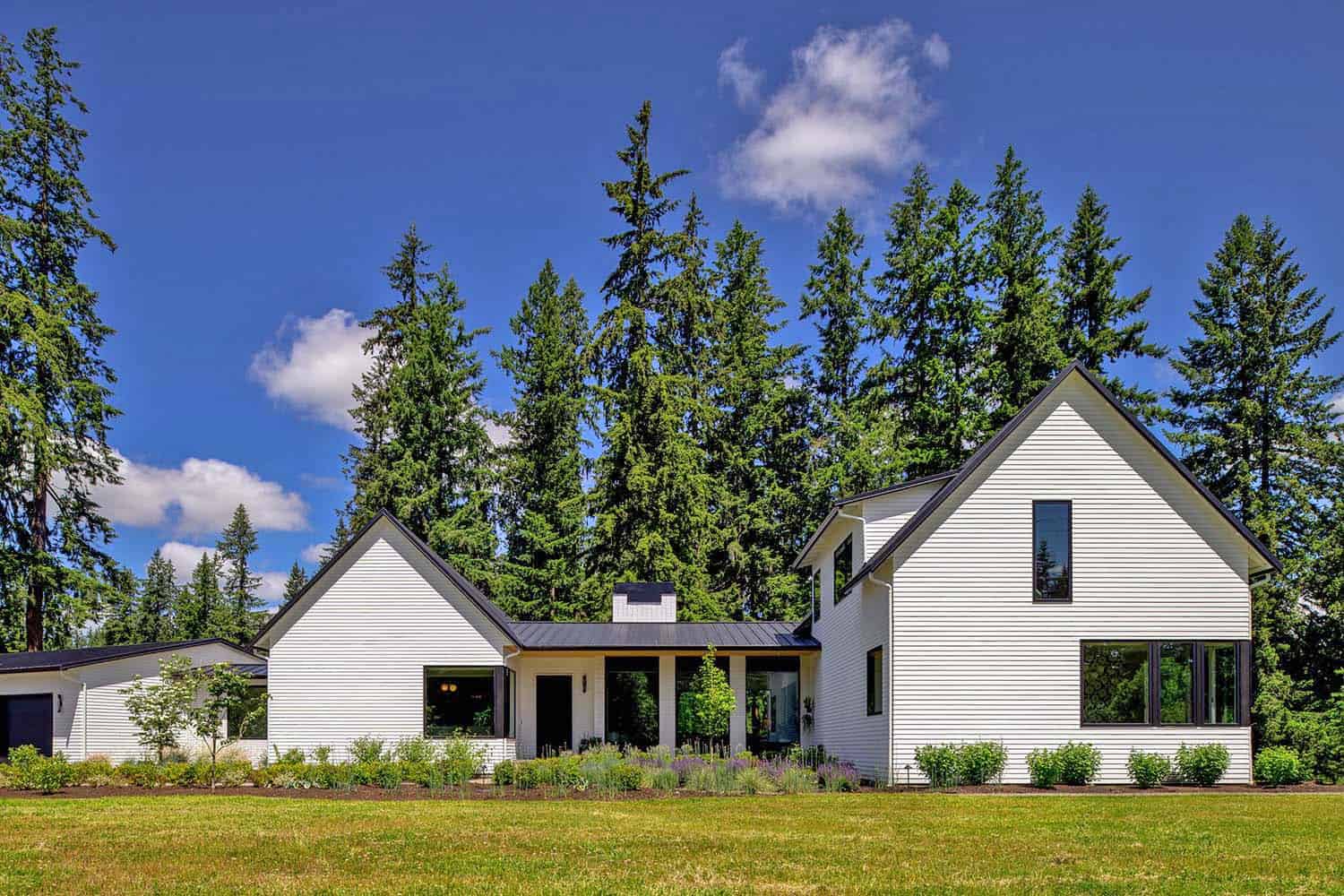 Modern white Pacific Northwest farmhouse with gabled roof surrounded by tall evergreen trees and green lawn