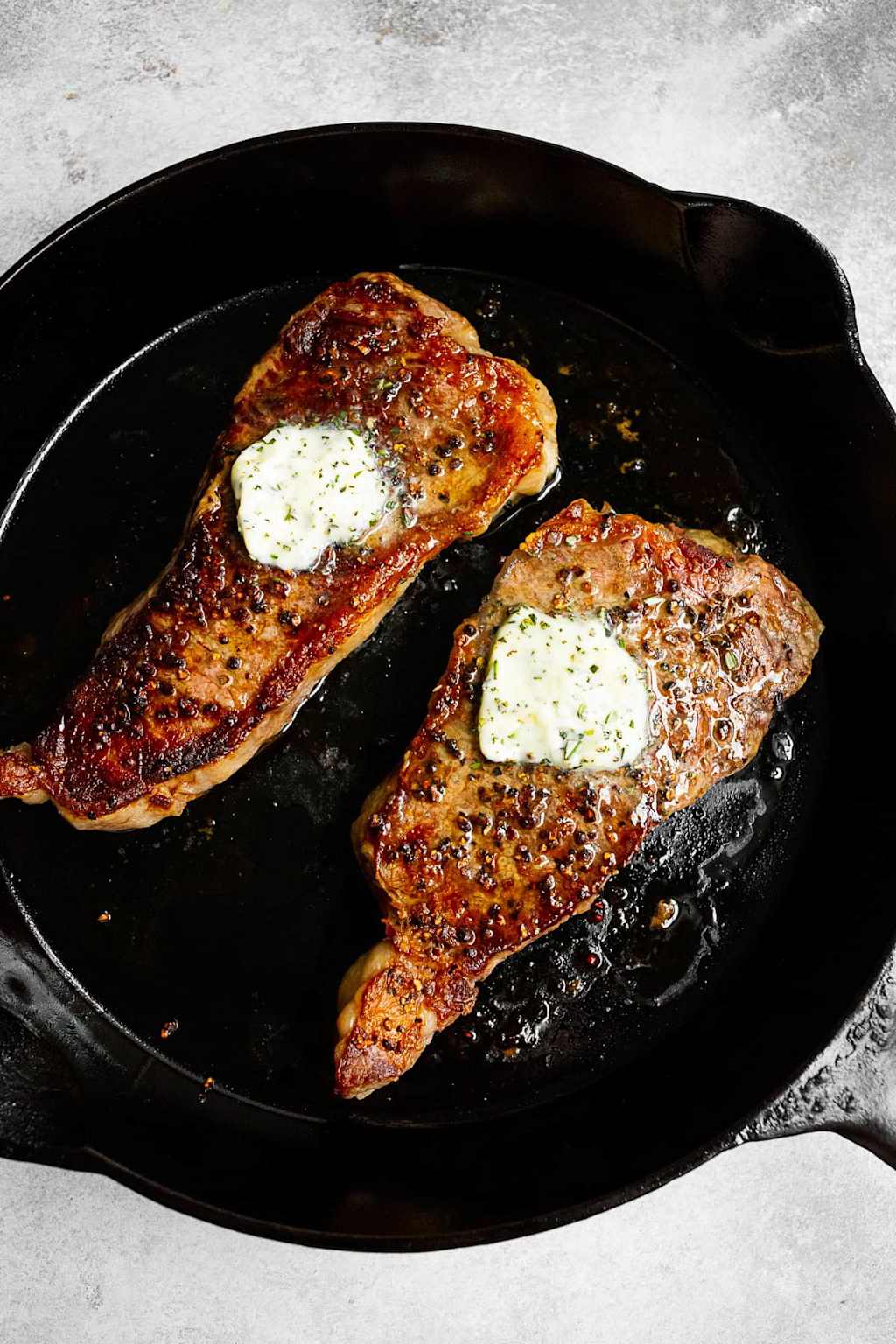 Two seared strip steaks with herb butter in a cast iron skillet