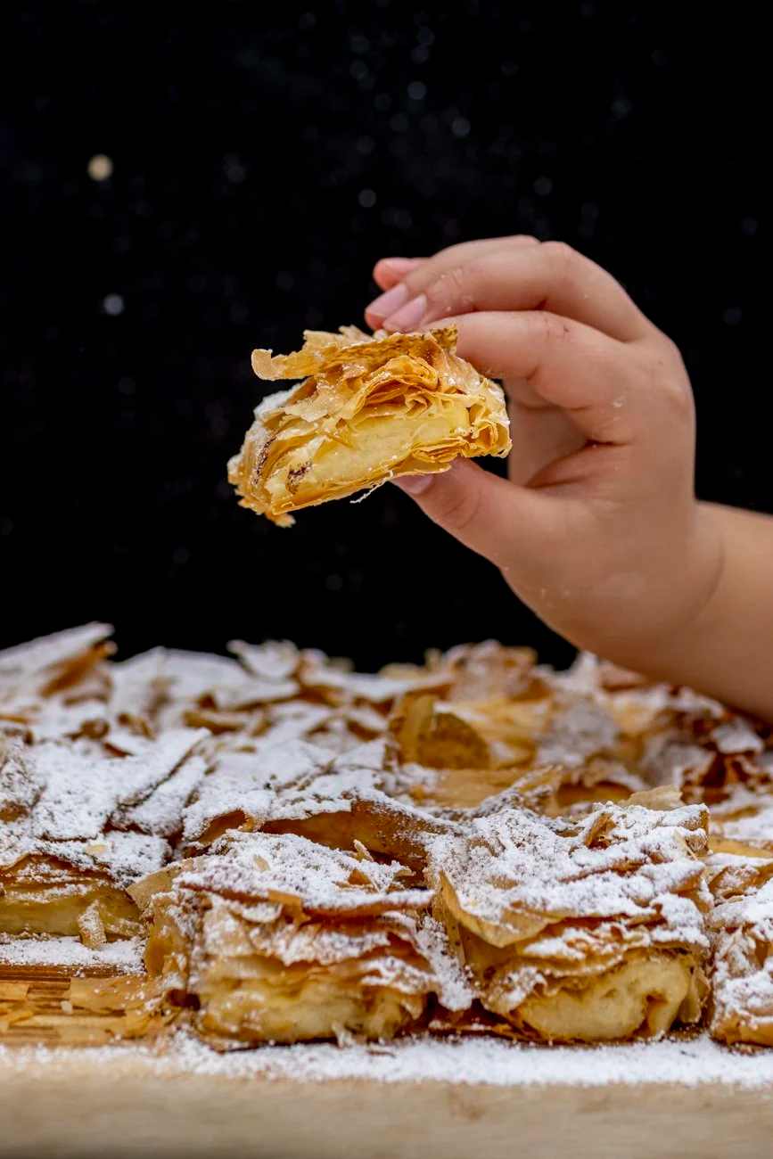Hand holding a piece of flaky bougatsa custard pastry with powdered sugar