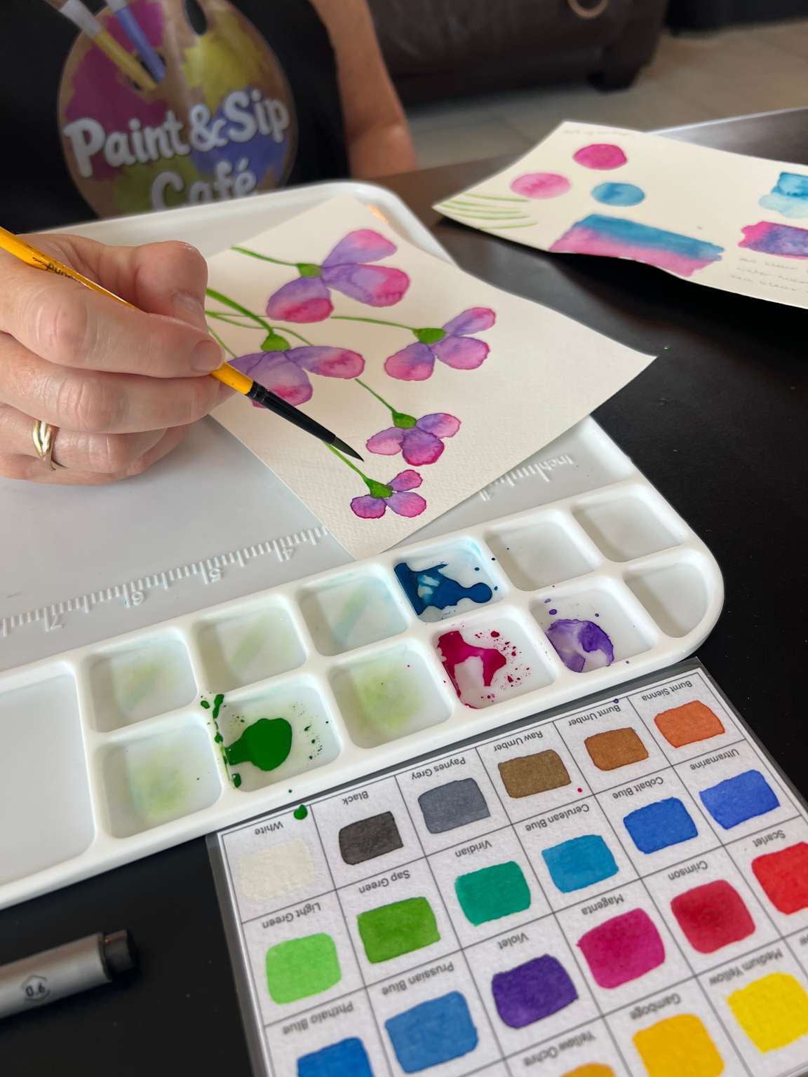 A hand painting purple flowers on paper using a brush with colorful watercolors nearby