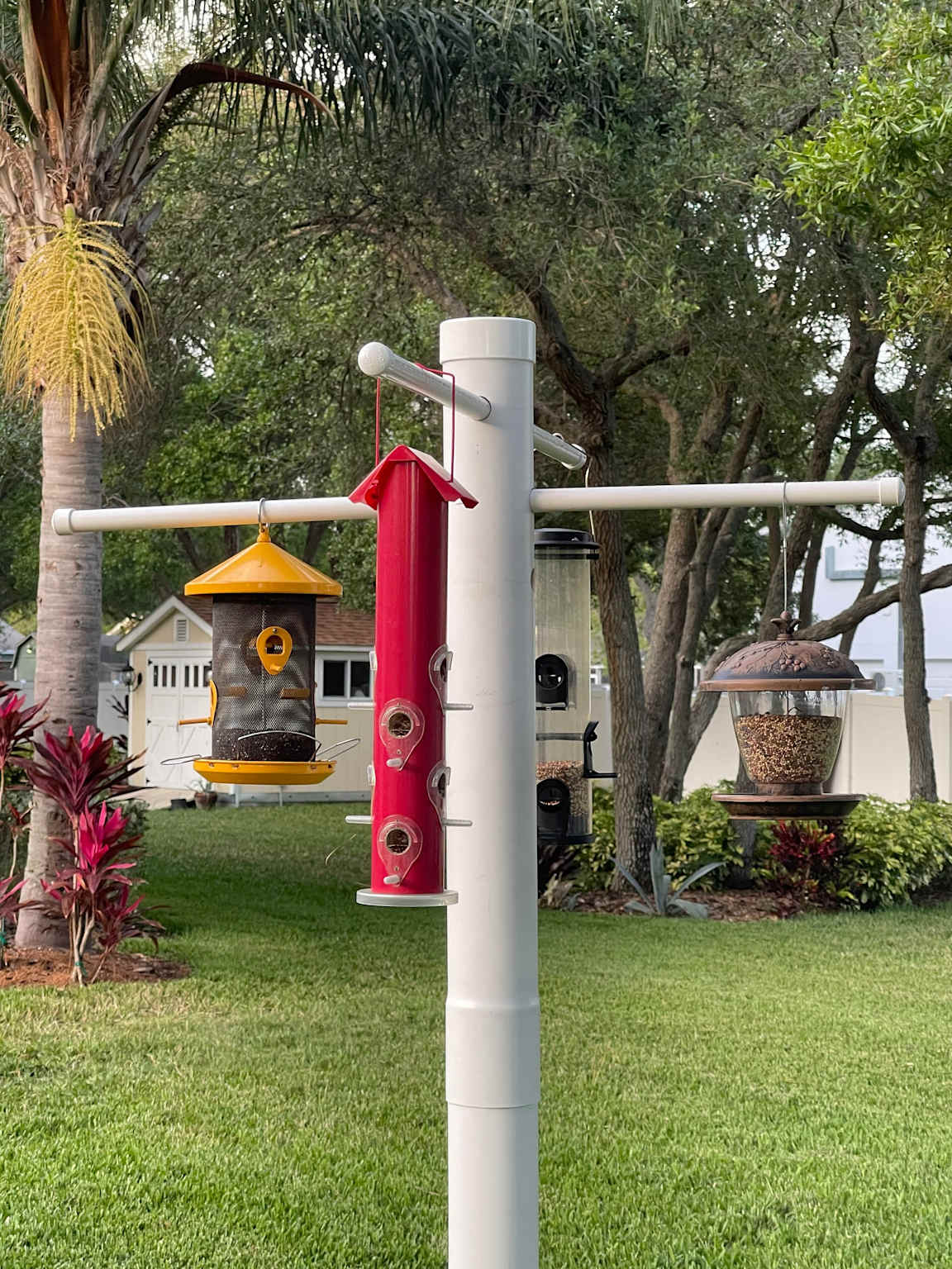 A white metal bird feeder pole system in an open backyard garden with multiple feeders