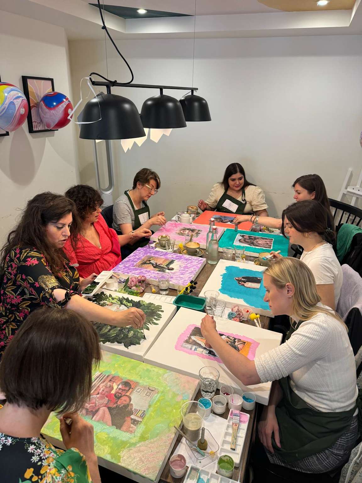 Group painting colorful artworks on canvases at a cafe table during a Paint and Sip workshop