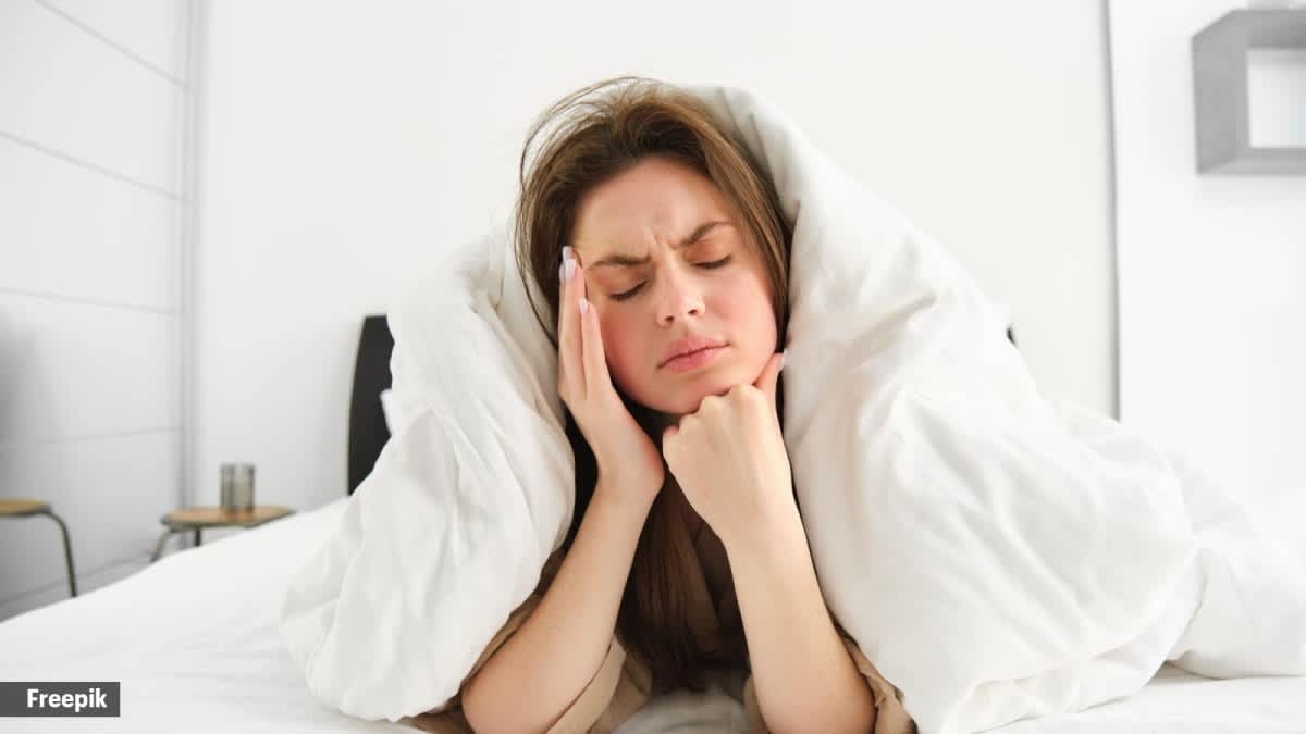 Person lying in bed with dishevelled hair holding forehead with fatigued expression — illustrating the non-restorative sleep quality experienced during fever and illness despite spending extended hours in bed