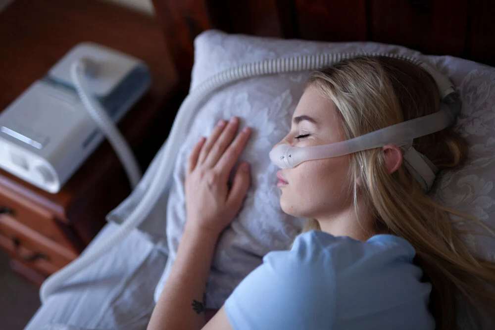 Woman sleeping in bed wearing a CPAP nasal mask connected by tube to a white CPAP machine on the bedside table — showing sleep apnea treatment in a real bedroom setting