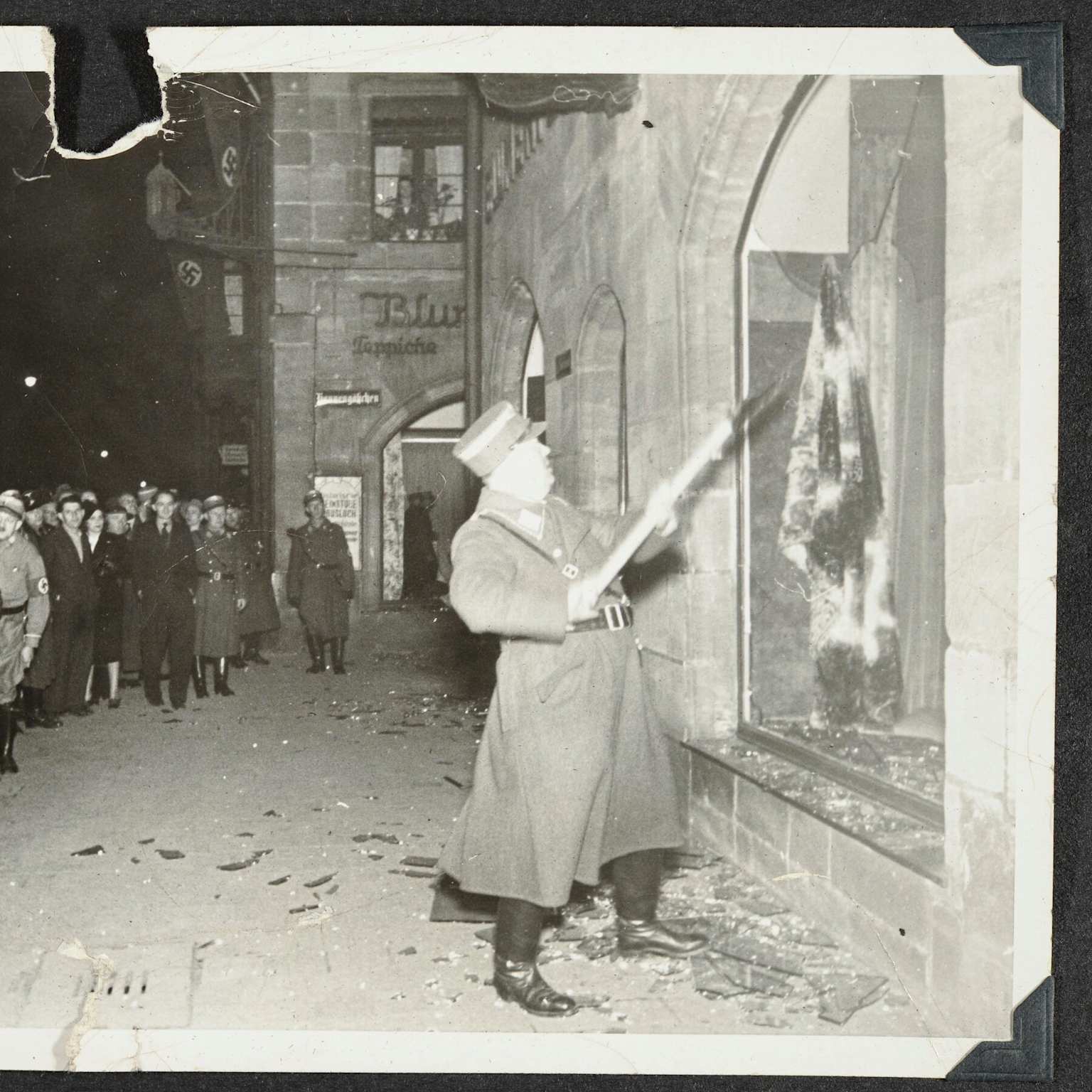 A Nazi officer smashes the window of a Jewish-owned shop on Kristallnacht, November 9-10, 1938, as crowds watch.