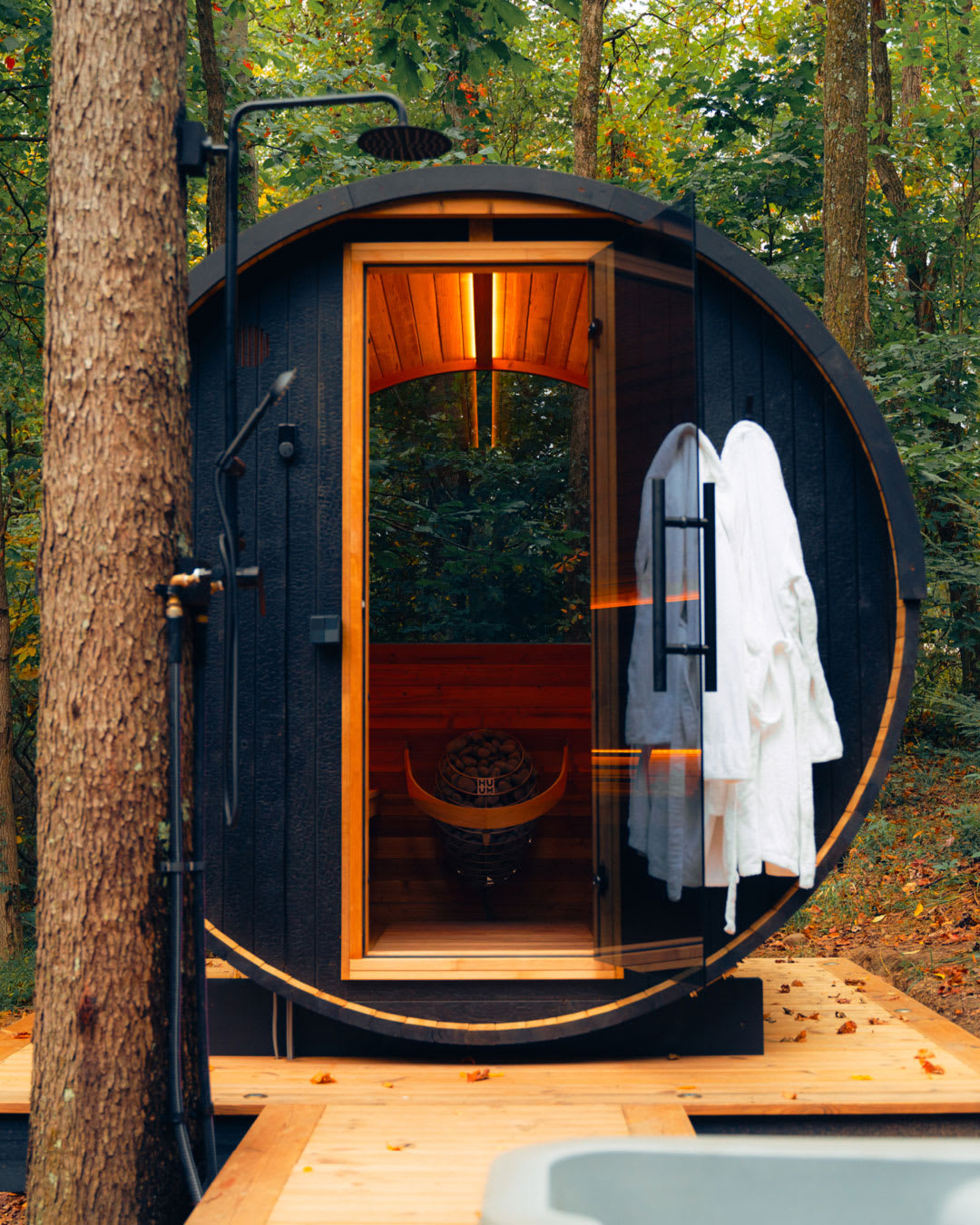 Outdoor barrel sauna with open door showing interior benches and heater, with an outdoor shower and wooden deck alongside