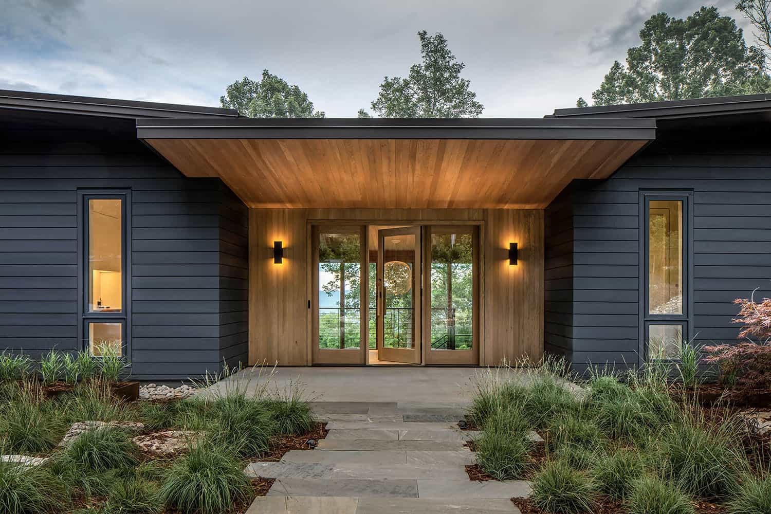 Modern mountain home exterior with open front door, dark siding, wood entry, and surrounding trees