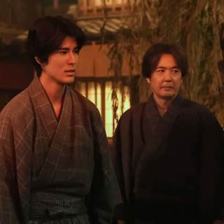 Two men in traditional kimono standing in a warmly lit old Japanese street scene, likely from NHK drama 'ばけばけ'