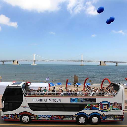 An open-top double-decker Busan City Tour bus on a coastal road with the Busan Harbor Bridge.