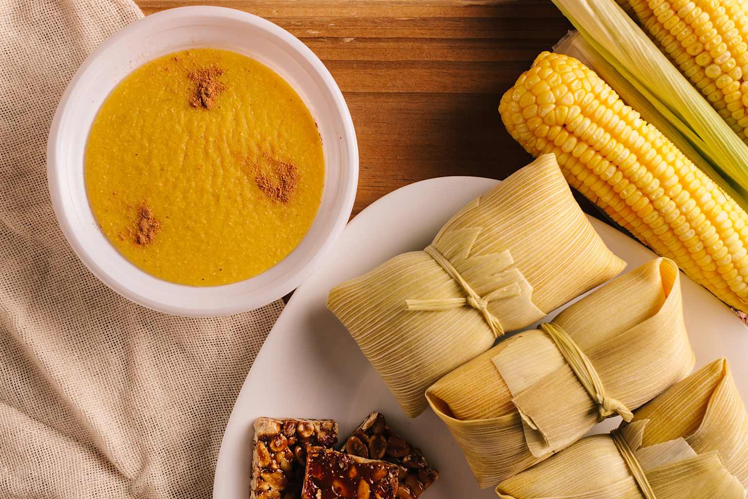 A collection of traditional Festa Junina treats, including pamonha, curau or canjica, fresh corn, and pé de moleque