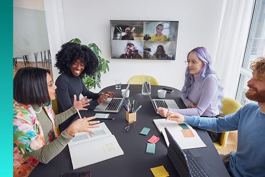 Hybrid work meeting with in-person and remote team members collaborating using laptops and video conferencing.