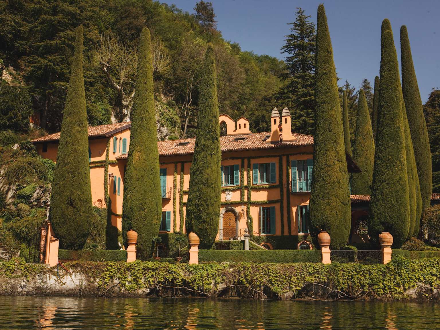 A grand, peach-colored villa surrounded by tall cypress trees, situated directly on the serene waters of a lake, resembling properties found in Lake Como