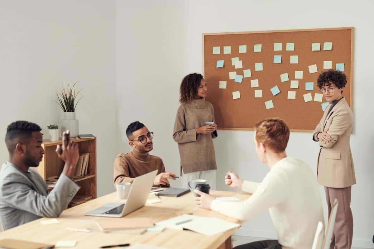 A diverse team engaged in a collaborative meeting with brainstorming sticky notes on a corkboard, illustrating employee engagement and inclusive workplace culture.