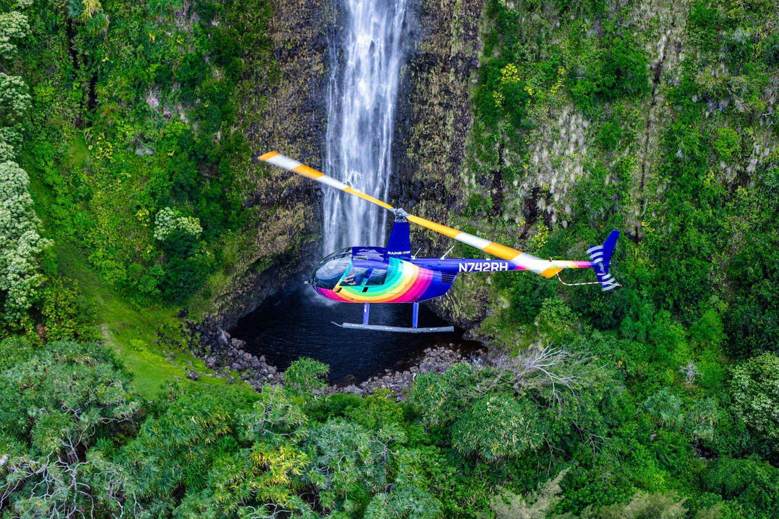A colorful helicopter offers breathtaking aerial views of a majestic Hawaiian waterfall amidst verdant landscapes