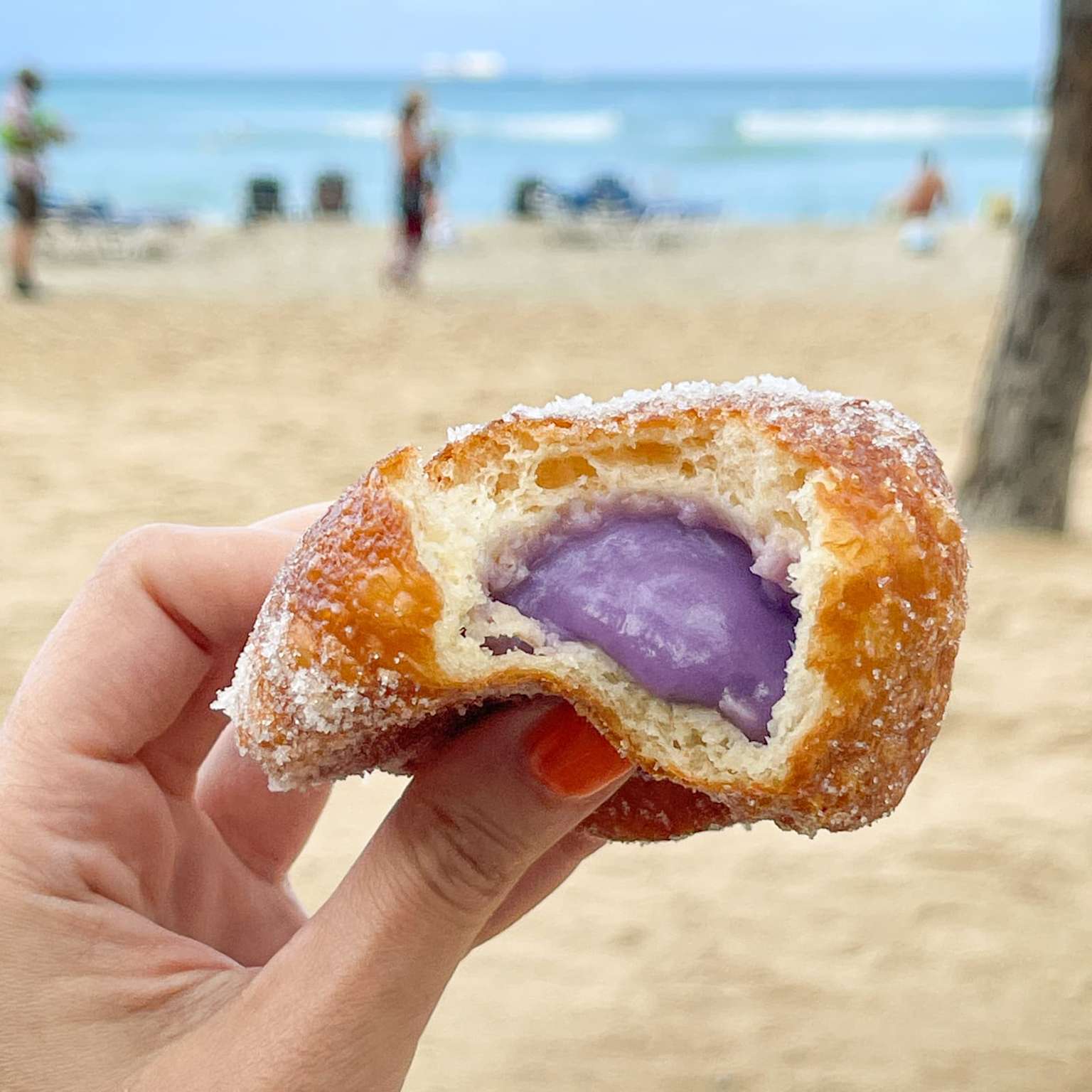 A hand holding a sugar-coated malasada with purple filling at a Hawaiian beach, showcasing a popular local treat from Leonard's Bakery