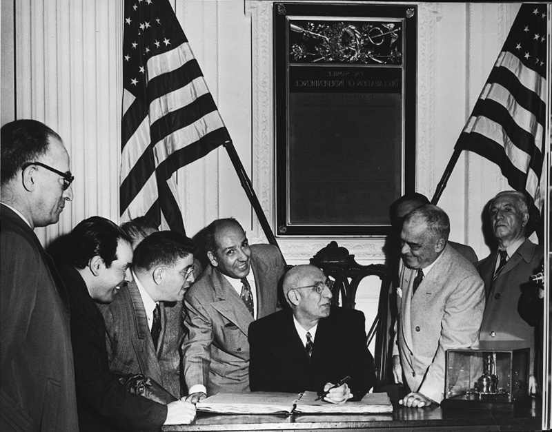 Prime Minister Mohammad Mossadegh seated at a desk, surrounded by other officials, with American flags in the background