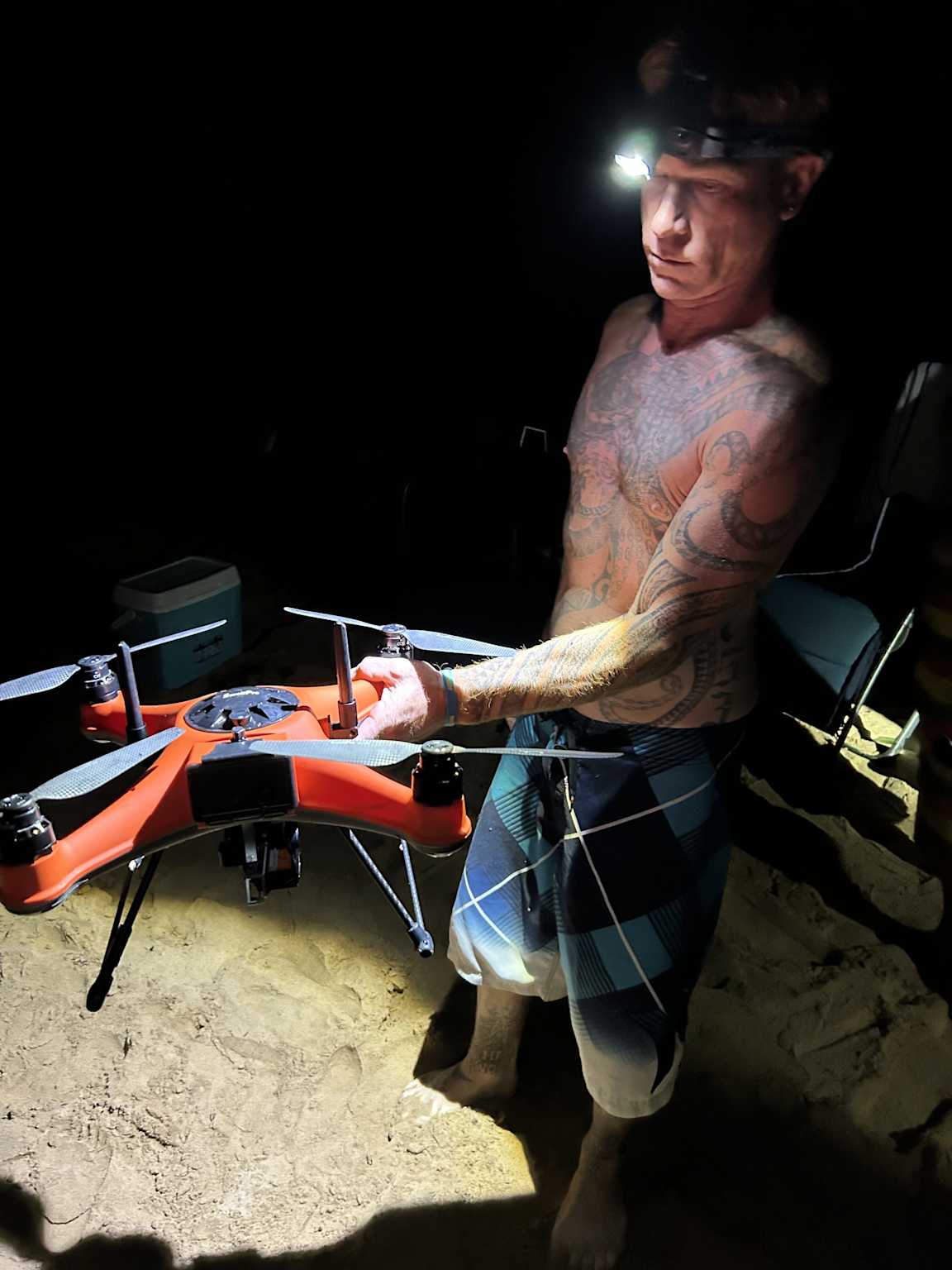 A man with a headlamp holds a waterproof drone on a sandy beach at night