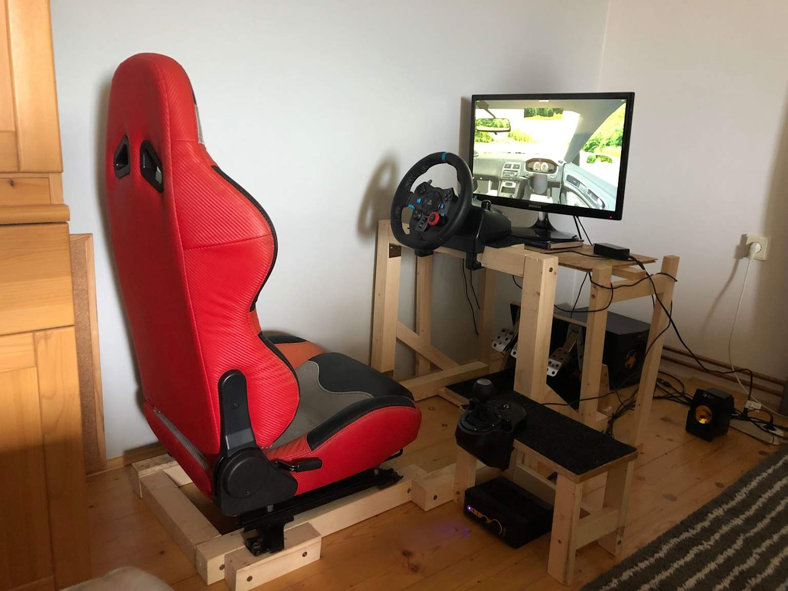 A DIY sim racing setup featuring a red racing seat and a Logitech G29 steering wheel with pedals mounted on a wooden frame