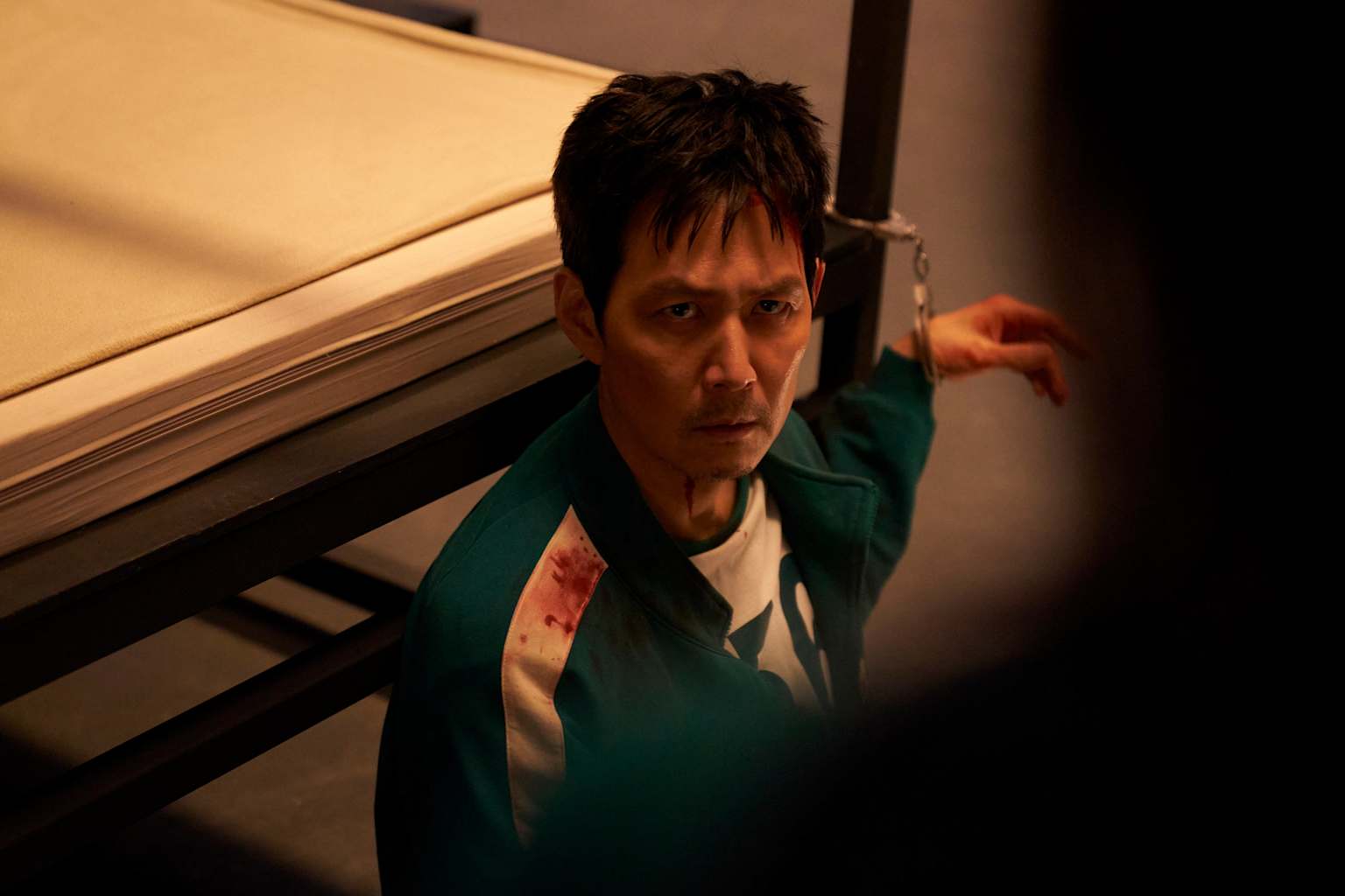 Gi-hun from Squid Game appears distressed and cuffed, with bloodstains on his tracksuit.