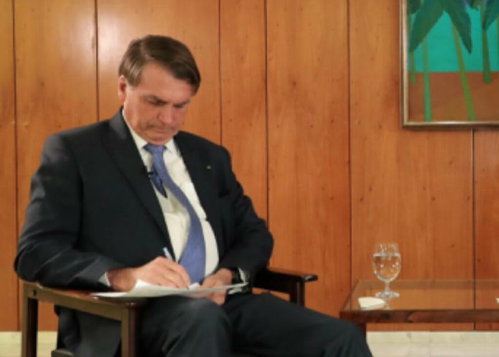 Jair Bolsonaro appearing to sign a document while seated in a formal setting.