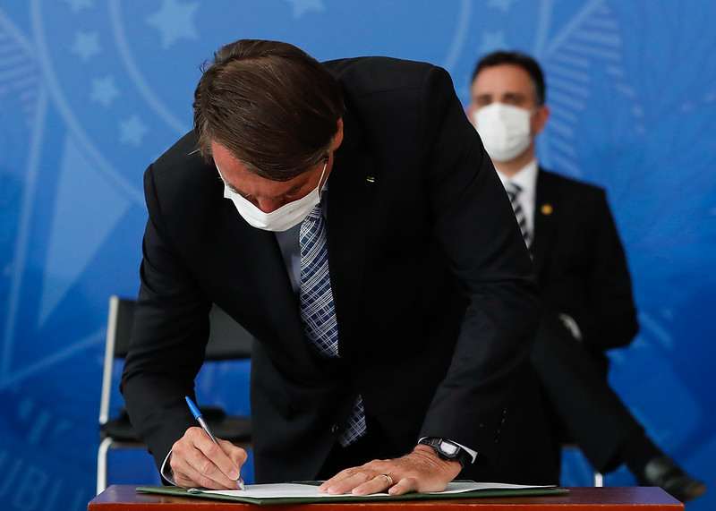 Jair Bolsonaro signs a document while wearing a face mask in an official setting.