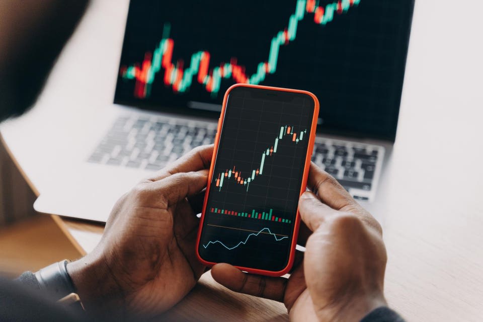 A person analyzes stock market data on a smartphone, with a laptop displaying similar charts in the background, illustrating the act of monitoring investments