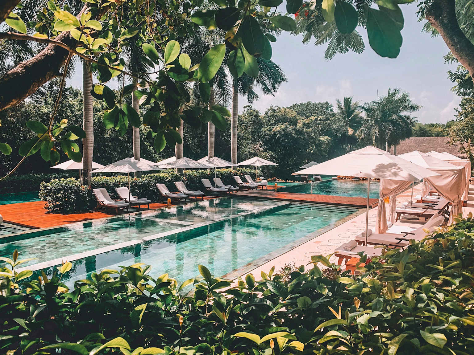 A serene, luxurious resort pool area surrounded by lush tropical greenery, featuring multiple pools and comfortable lounge seating