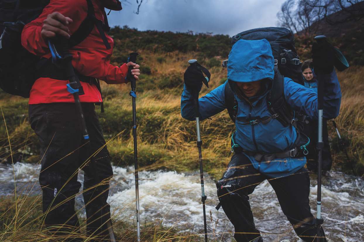 Hikers crossing a fast-flowing stream in wet, challenging mountain weather, highlighting the need for caution and proper gear