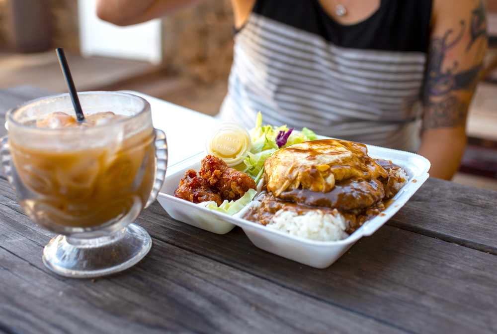 A classic Hawaiian plate lunch with rice, hamburger patty, fried egg, fried chicken, and salad served with iced coffee at a casual outdoor setting