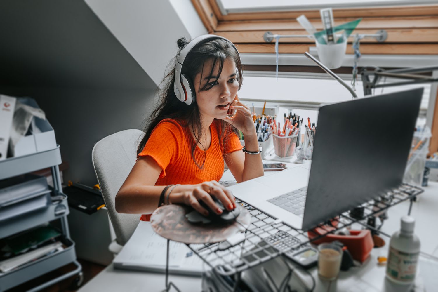 Young adult wearing headphones and working at a cluttered desk, focused on a laptop screen in a bright room
