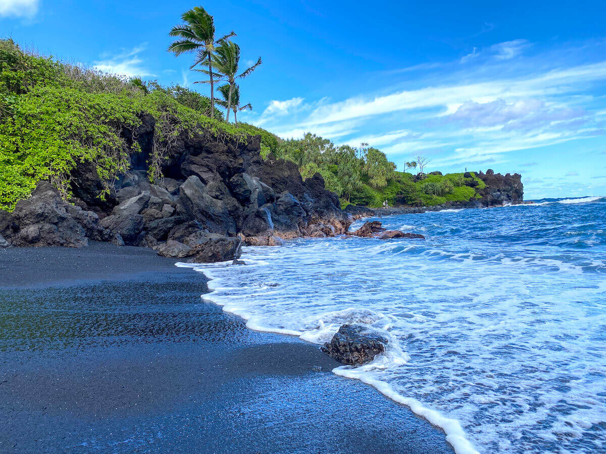 Black sand beach with lush greenery and volcanic cliffs along the coast of Hana, Maui