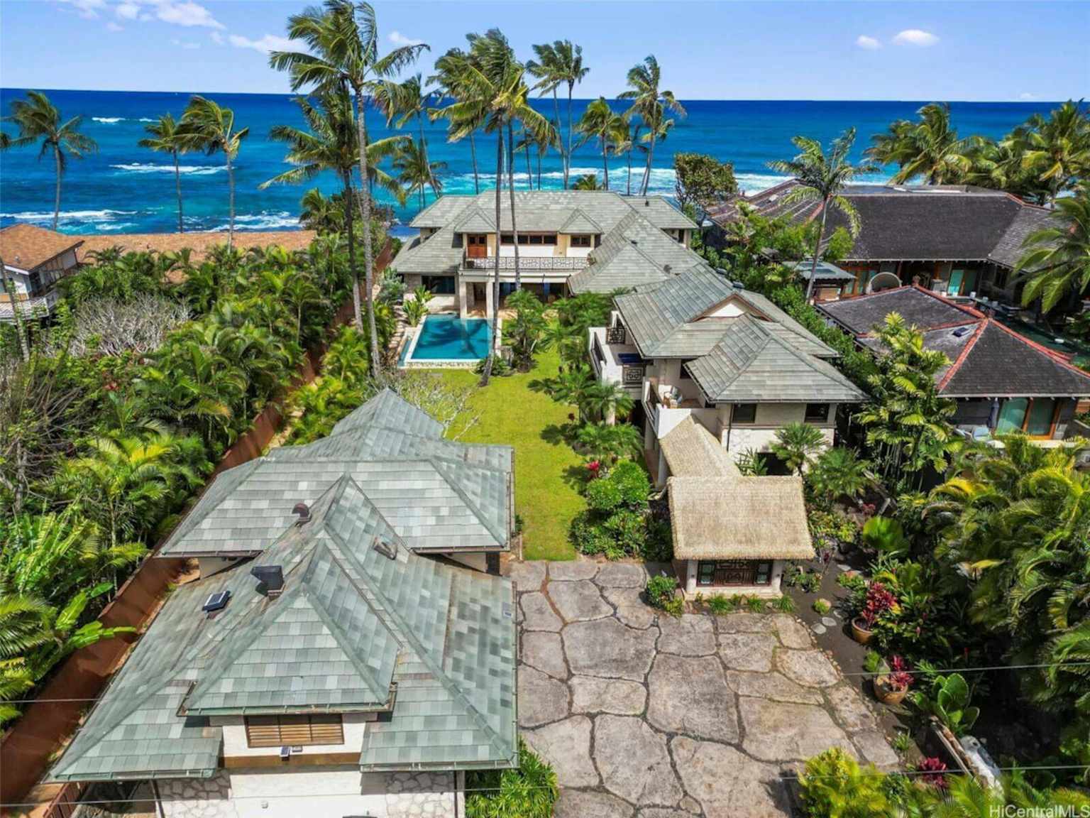 An aerial view of a luxurious North Shore Oahu oceanfront estate with palm trees, swimming pool, and modest neighboring homes near Sunset Beach
