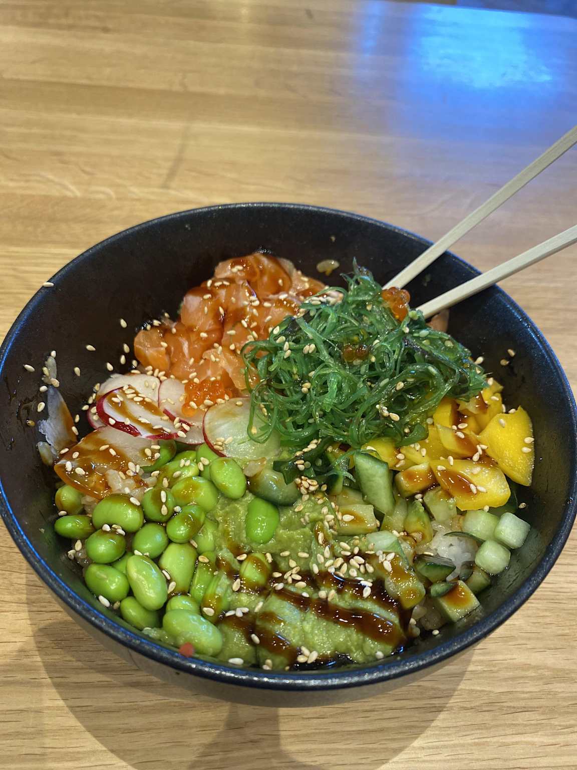 A fresh Hawaiian poke bowl featuring salmon, seaweed salad, edamame, mango, and sesame seeds ready to be enjoyed with chopsticks