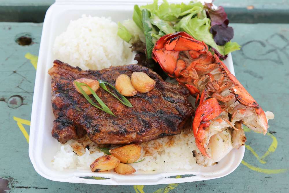 Hawaiian surf and turf meal with grilled steak, lobster tail, rice, and greens served in a casual takeout container