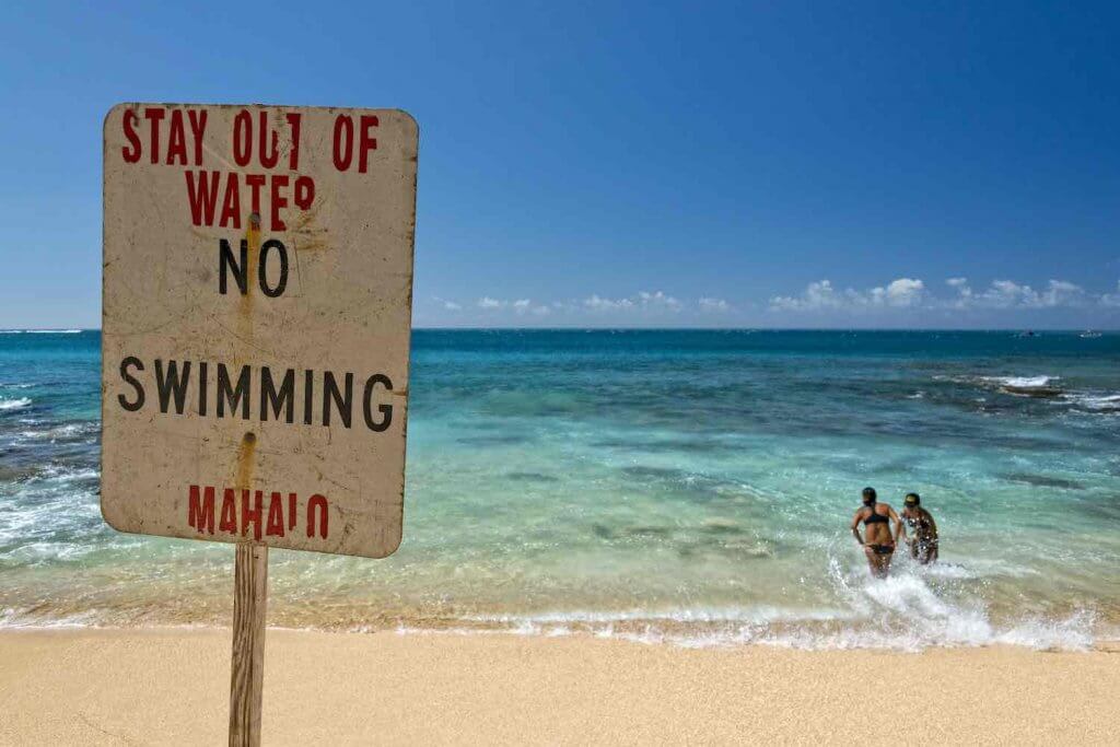 A worn warning sign on a Hawaiian beach instructing no swimming due to ocean dangers, highlighting a common tourist mistake of ignoring safety advice