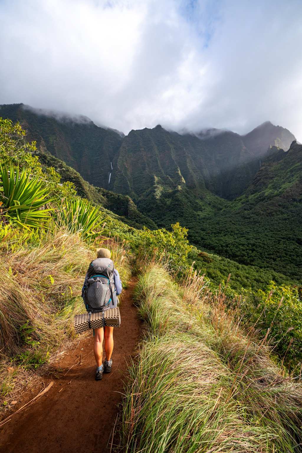 A hiker backpacks along the narrow Kalalau Trail on Kauaʻi's Nāpali Coast surrounded by dramatic mountain scenery and misty clouds
