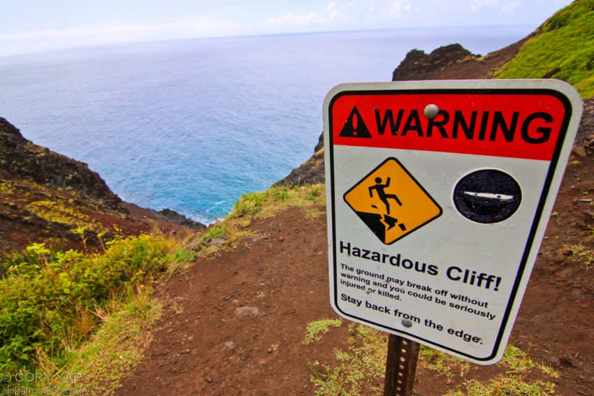 Warning sign indicating hazardous cliff conditions on a Hawaii coastal hiking trail with ocean view in the background