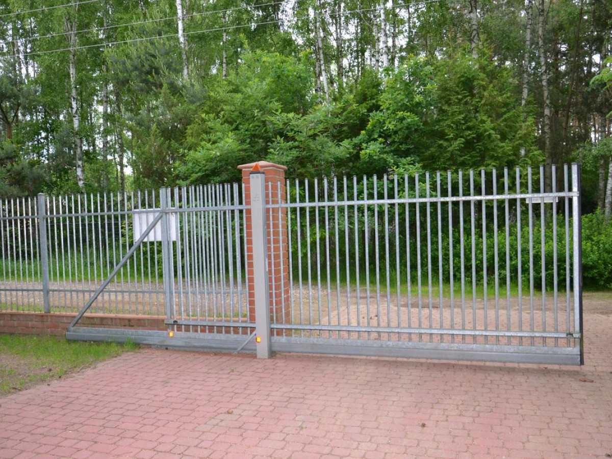 Modern silver sliding gate installed on a paved driveway beside a green wooded area, demonstrating the design and functionality of a cantilever sliding gate