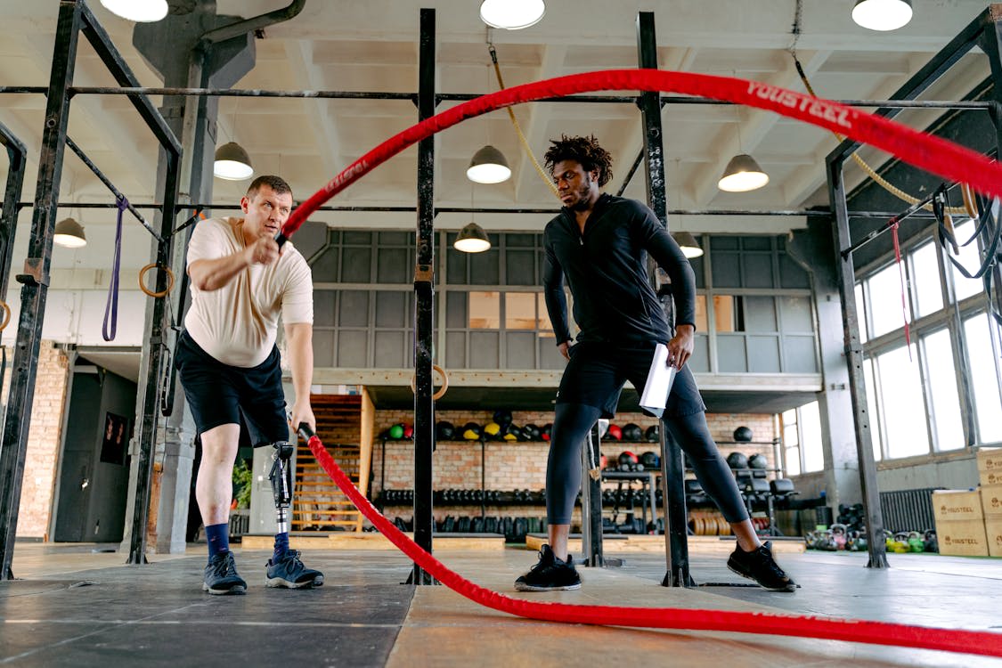 A personal trainer guides a man with a prosthetic leg through an adaptive workout using battle ropes, showing the benefits of personalized fitness training for people with disabilities