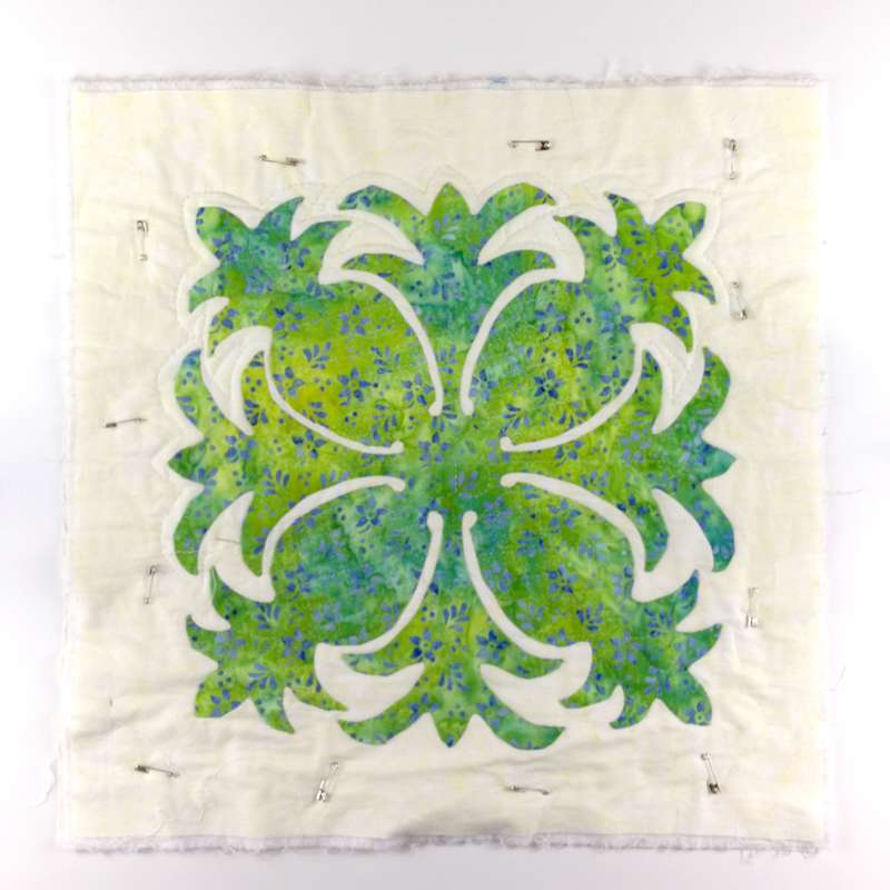 Traditional Hawaiian quilt square with intricate green and blue floral applique pattern on cream background in progress