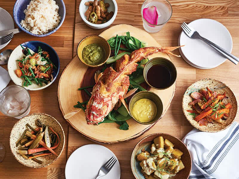 Family-style Hawaiian meal featuring whole fish, varied side dishes, and communal sharing on rustic table