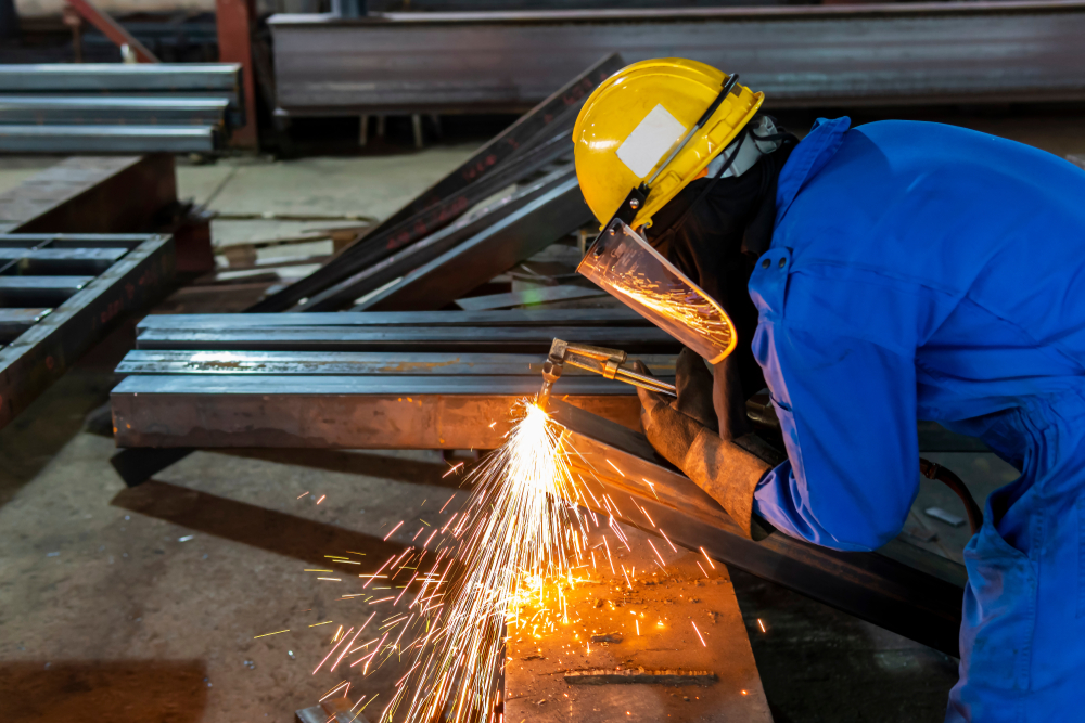 A worker uses a cutting torch to shape structural steel beams during fabrication, wearing full protective gear