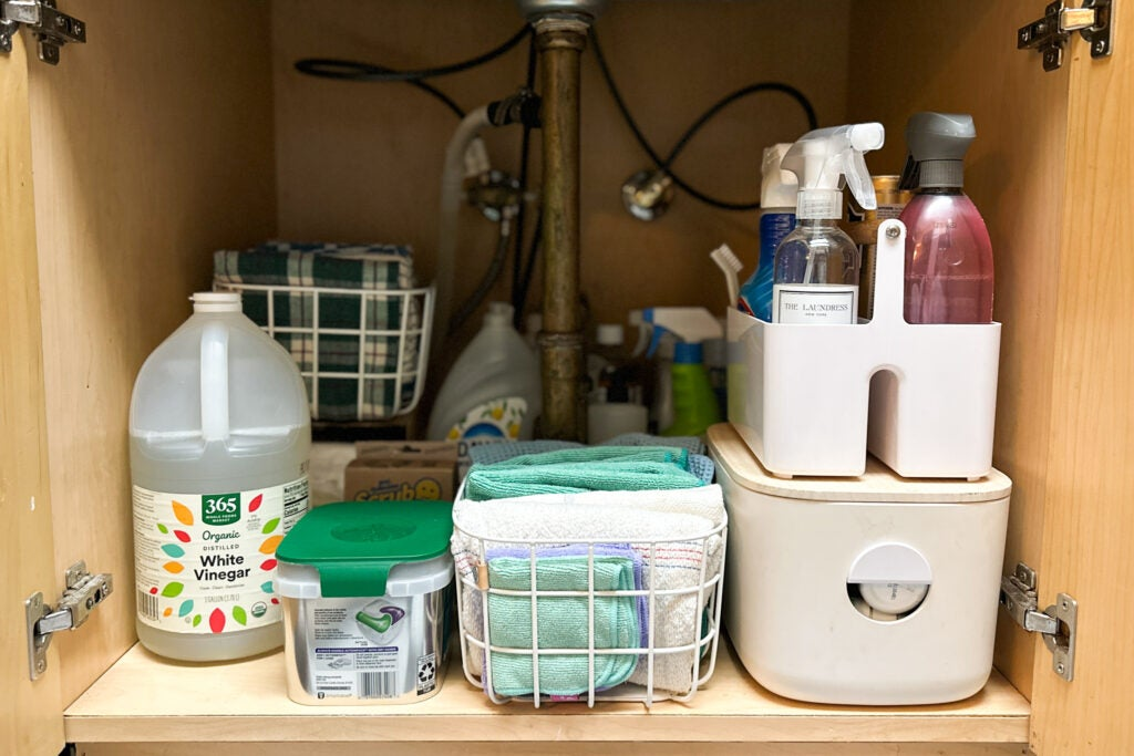 Organized and essential home cleaning supplies stored neatly under the sink, including white vinegar, detergent pods, microfiber cloths, and cleaning sprays