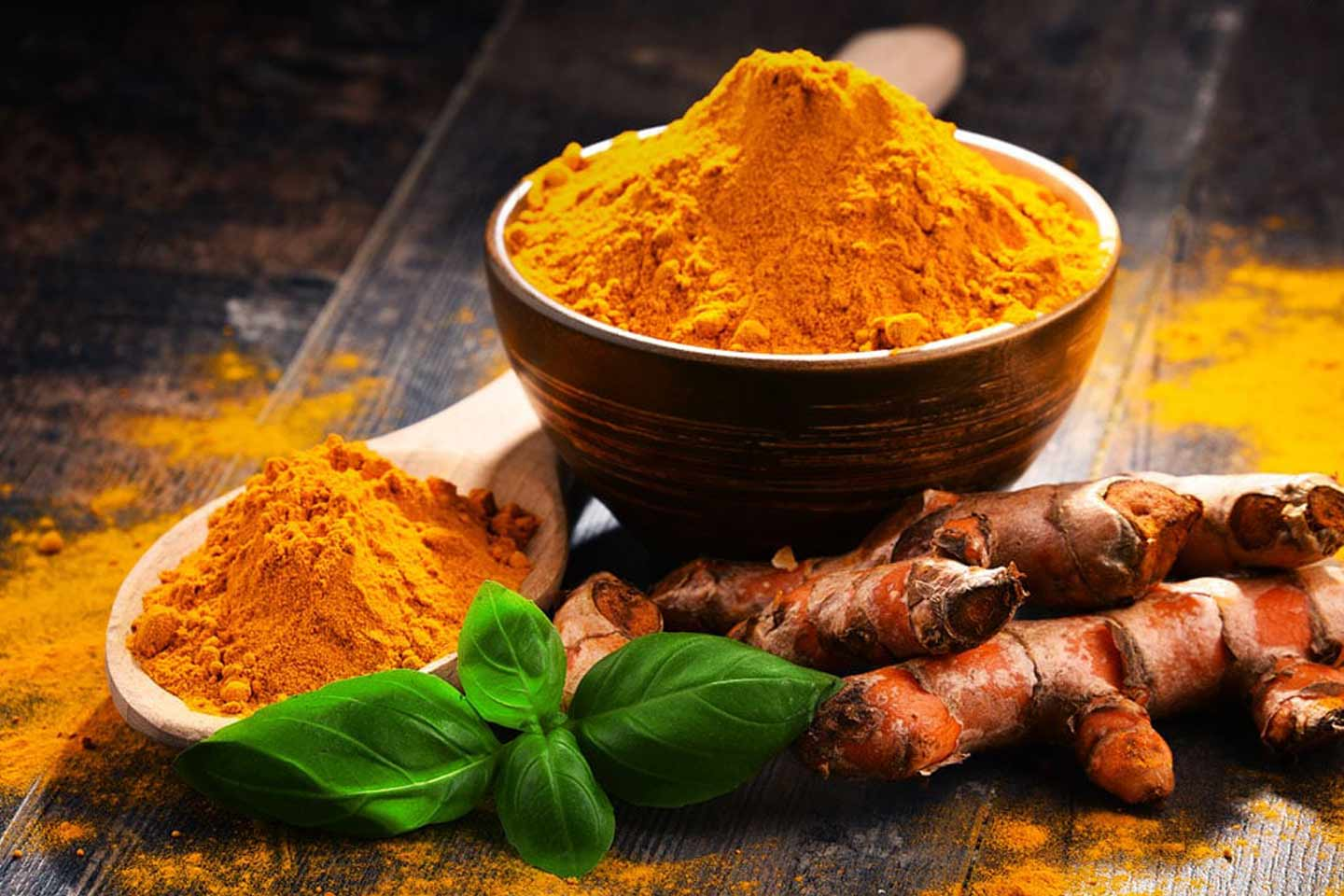 Fresh turmeric roots and bright turmeric powder displayed with green basil leaves on a rustic wooden table