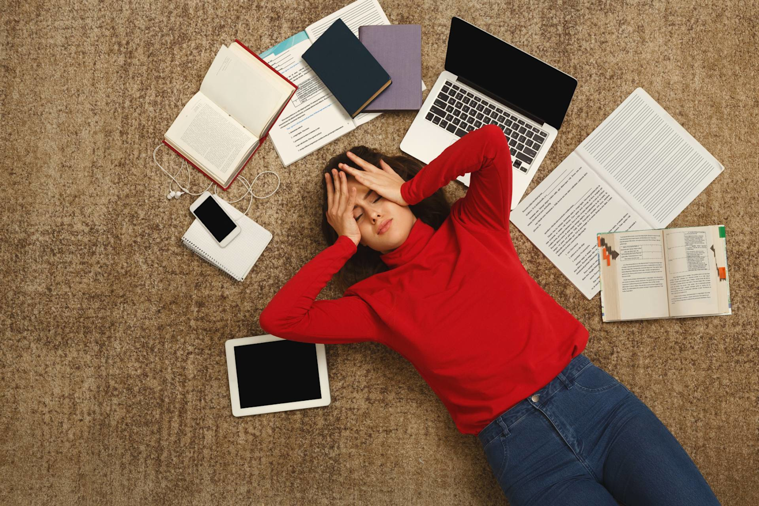 A student lies overwhelmed by academic pressure amid study materials and devices