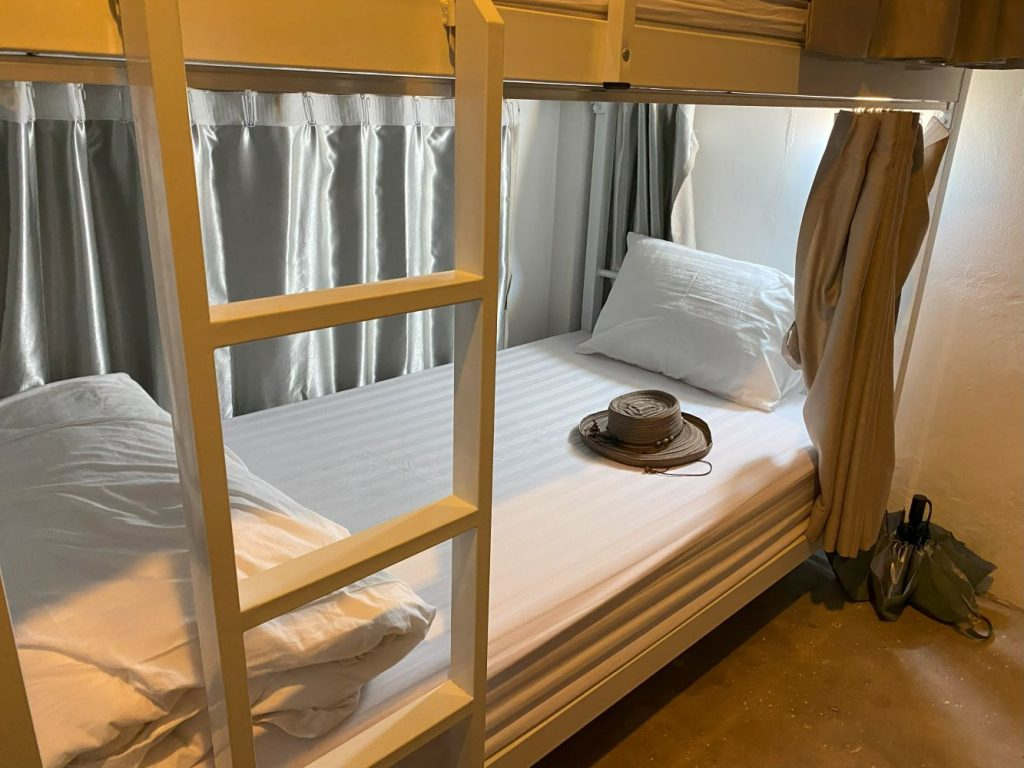 A neatly made lower bunk bed in a budget hostel dormitory in Southeast Asia with privacy curtains and traveler's hat