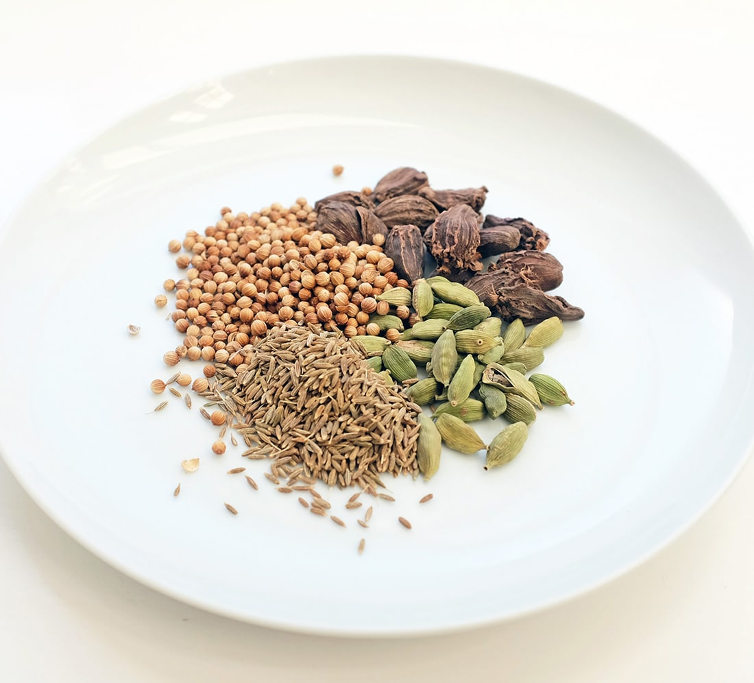 Coriander seeds, cumin seeds, green cardamom, and black cardamom pods on a white plate, commonly used low FODMAP Indian spices
