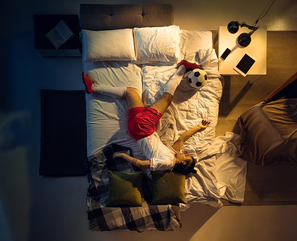 Teenage soccer player resting in bed with a soccer ball, illustrating sleep's role in athletic recovery and injury prevention
